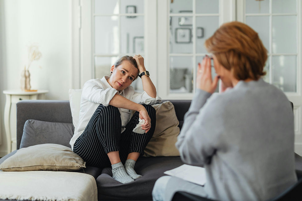 Therapist giving advice to anxious woman; one listens with concern, the other gestures with hands. Warm tones, cushions on couch, light streaming in.
