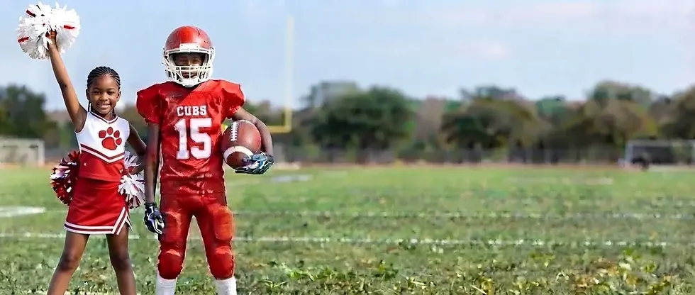 A child in a red football uniform holds a football on a green field. A goalpost is visible in the background under a clear sky.