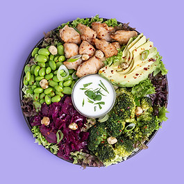 Protein Salad. Ein schwarzer Teller auf lila Untergrund, gefüllt mit Blattsalat, Brokkoli, Avocado, Edamame, Hühnchen, Rotkohlsalat und einem Joghurtdressing.
