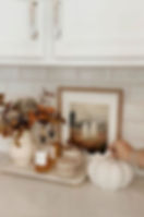 Fall kitchen countertop vignette with white subway tile backsplash, white cabinets, a tray with caramel syrup bottles, cream mugs, a neutral vase with autumn foliage, framed ghost-themed fall art, and a white ceramic pumpkin.
