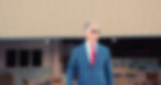 John Glen Weaver stands in a blue suit and red tie outside an aviation hangar.