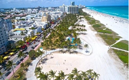 Turistas aproveitando a praia de Miami Beach em um dia ensolarado, com cadeiras de sol e mar azul ao fundo