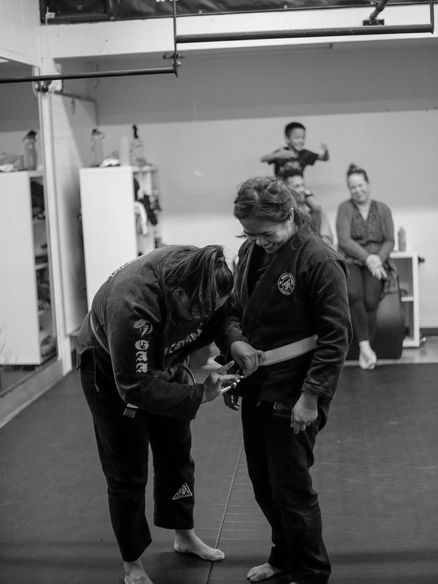 Women standing in gym with belt, getting stripes from female coach during grading.