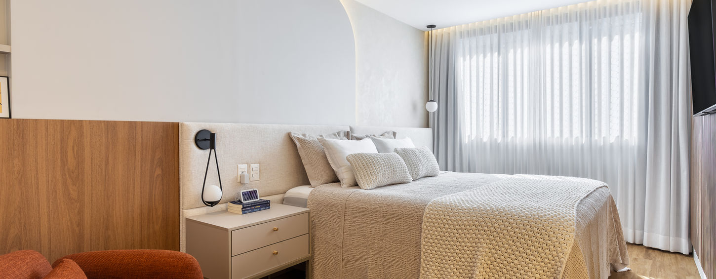 Quarto de casal de alto padrão com integração de marcenaria e estofaria. O ambiente apresenta painel de cama curvo com iluminação indireta, ladeado por painel amadeirado e mesa de cabeceira em laca fosca. Produção da marcenaria boutique TRACIATTO em Brasília, projetando o descanso através de uma estética atemporal.
