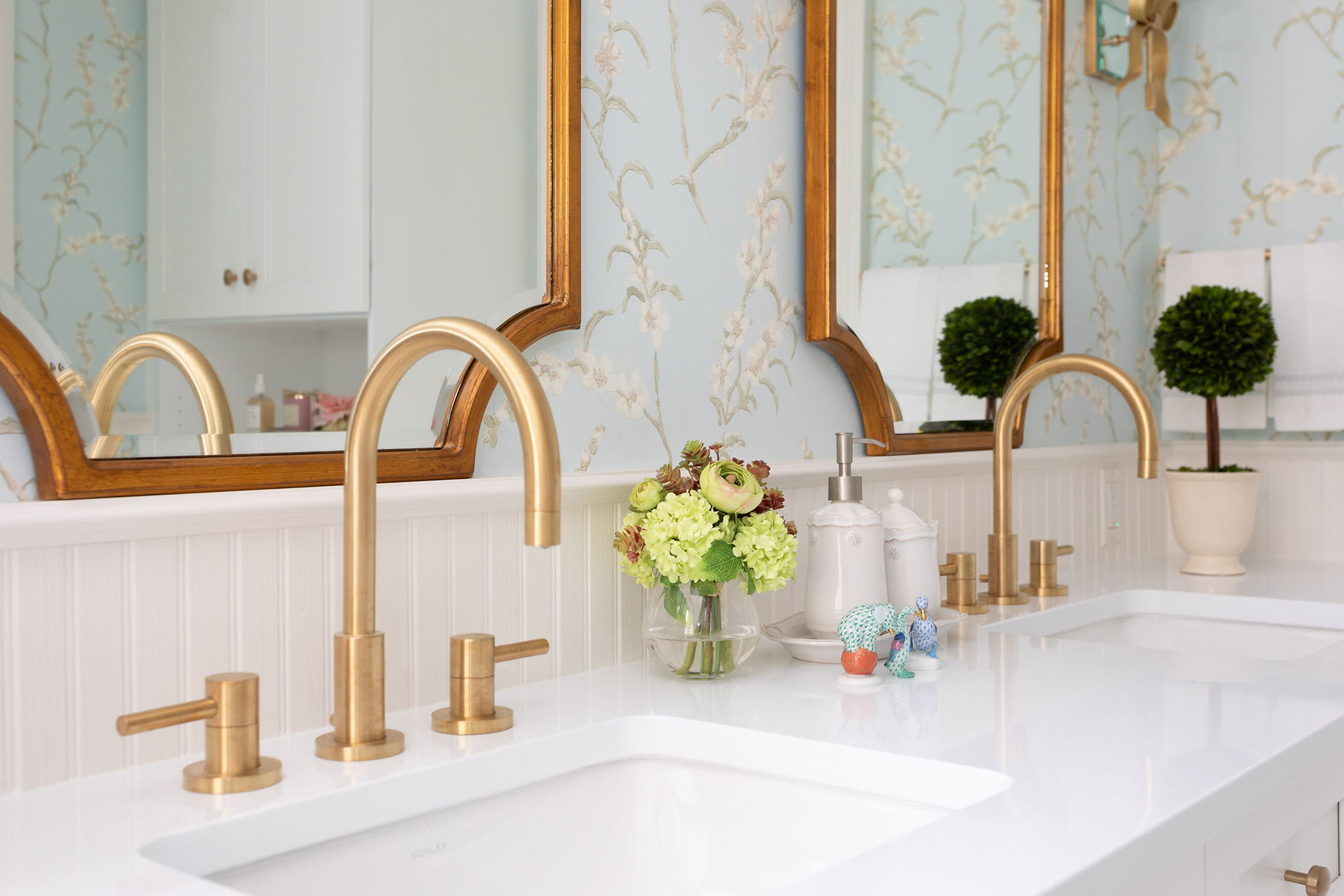 Brushed gold faucet with fresh flowers and soap dispenser on a white vanity counter beneath a wood-framed mirror.