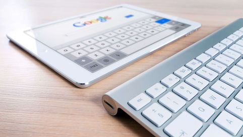 A modern tablet displaying the Google search engine logo next to a wireless keyboard on a wooden desk.