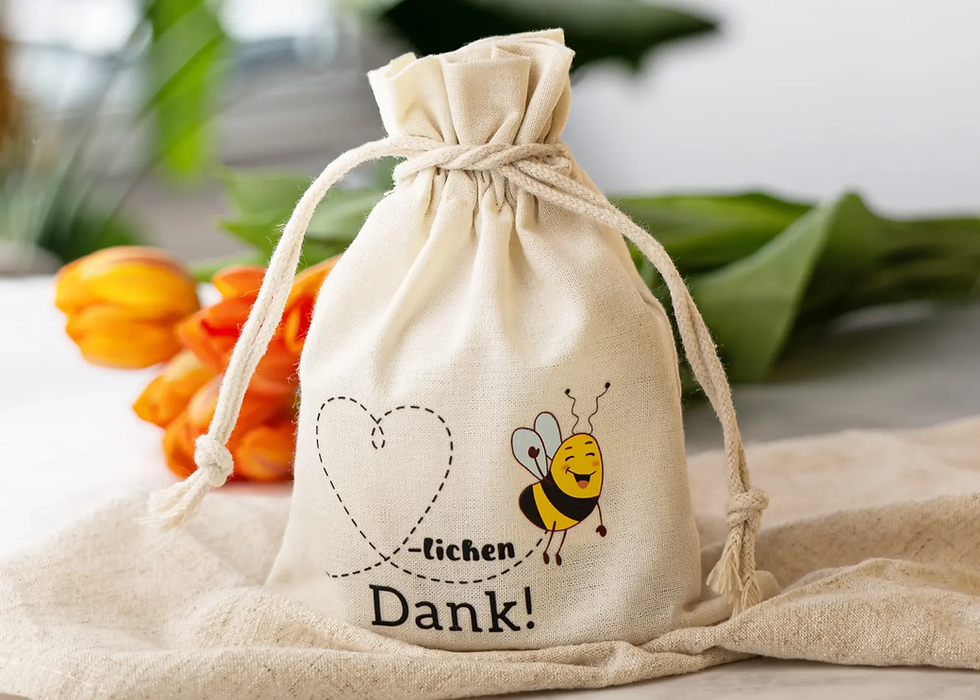 Stoffbeutel mit lachender Biene und Text "lichen Dank!" im Fokus. Im Hintergrund orange Tulpen, helle, fröhliche Atmosphäre.