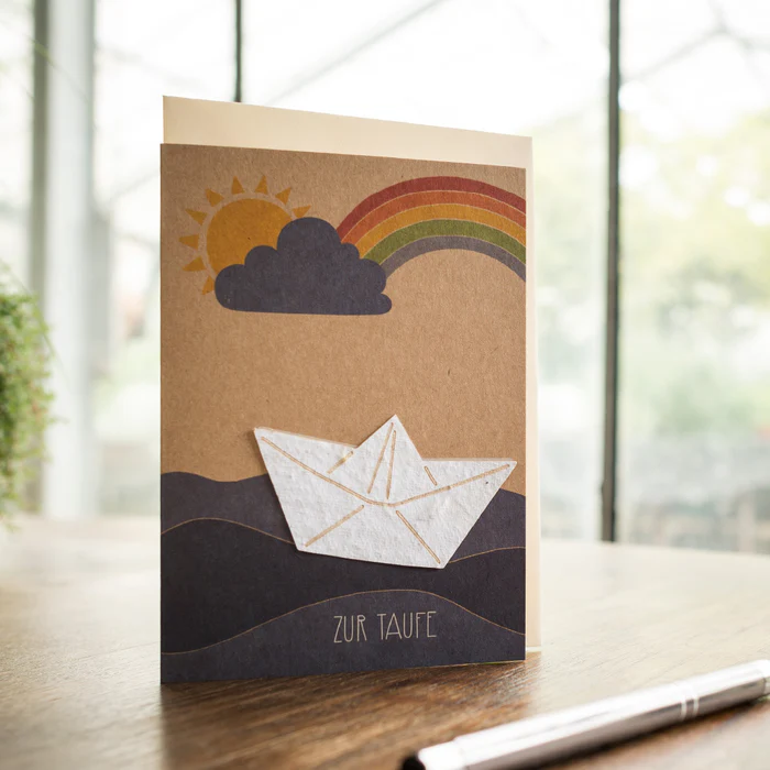Karte zur Taufe mit Papierboot, Sonne, Wolke und Regenbogen auf Holztisch, dazu der Text ZUR TAUFE.