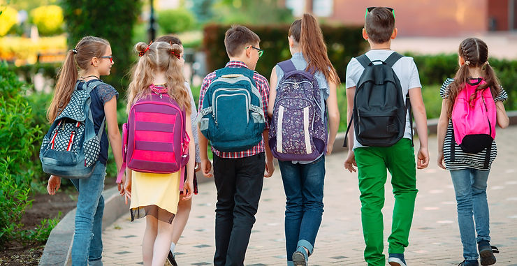 Sechs Kinder mit bunten Rucksäcken gehen auf einem sonnigen Schulweg davon.