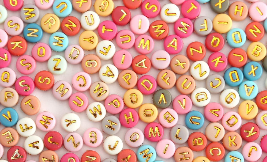 Bunte runde Buchstabenperlen in Pink, Gelb und Blau liegen dicht auf hellem Grund, als farbiges Muster.