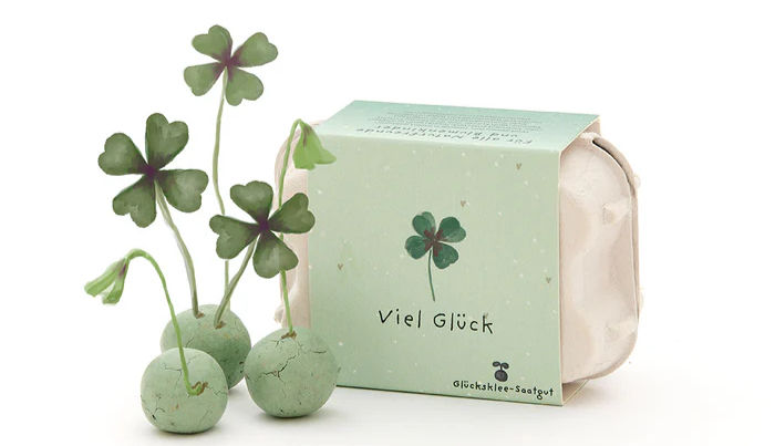 Verpackung mit vierblättrigem Glücksklee und grünen Saatkugeln, hellgrüne Box mit Aufdruck Viel Glück und Glücksklee-Saatgut