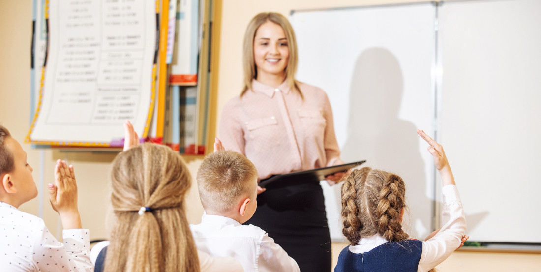 Lehrerin vor der Tafel im Klassenzimmer; mehrere Kinder sitzen an Tischen und melden sich fröhlich.