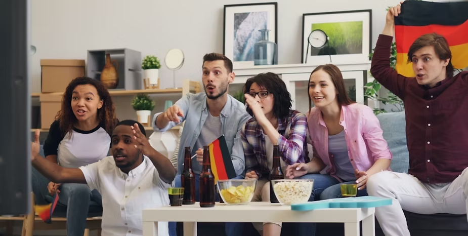 Gruppe jubelt vor Fernseher, hält Deutschlandfahne und Snacks. Wohnzimmer mit Bilderrahmen im Hintergrund, lebhafte Stimmung.
