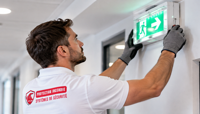 Technicien FPI2S en polo blanc avec logo rouge installant un bloc autonome d’éclairage de sécurité (BAES) au-dessus d’une porte.