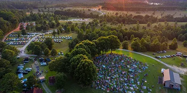 Aerial view of an outdoor concert with a crowd on a grassy field. Cars parked nearby, surrounded by trees. Warm sunset lighting.