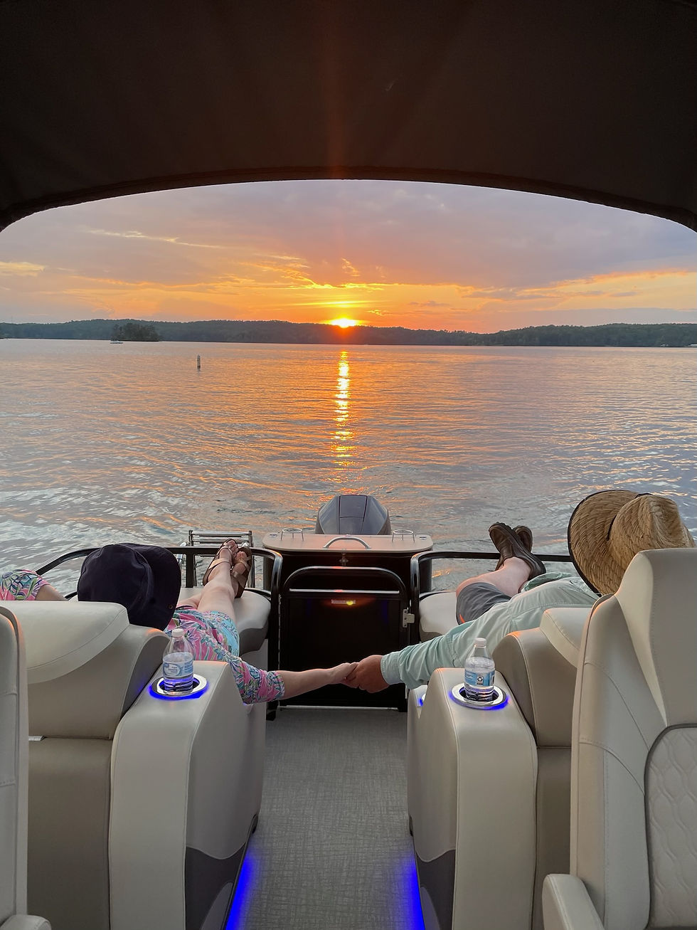 Two people relax on a boat, holding hands as the sun sets over a serene lake. Warm colors fill the sky, creating a peaceful mood.