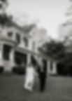 Couple in wedding attire holding hands, walking on lawn in front of a large house with columns. Smiling faces, trees in background. Black and white.