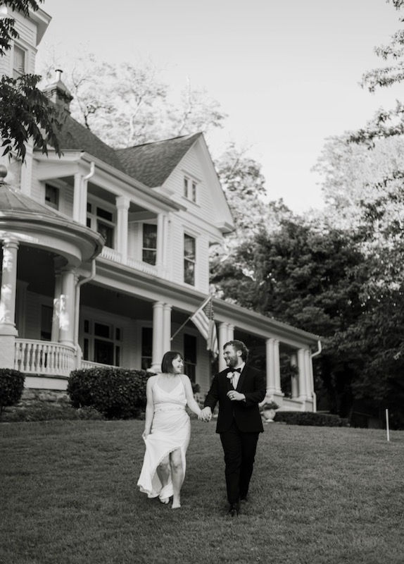 Couple in wedding attire holding hands, walking on lawn in front of a large house with columns. Smiling faces, trees in background. Black and white.