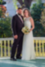 A couple in wedding attire smiles on a porch with a green and yellow railing. The bride holds a bouquet of white and pink flowers.