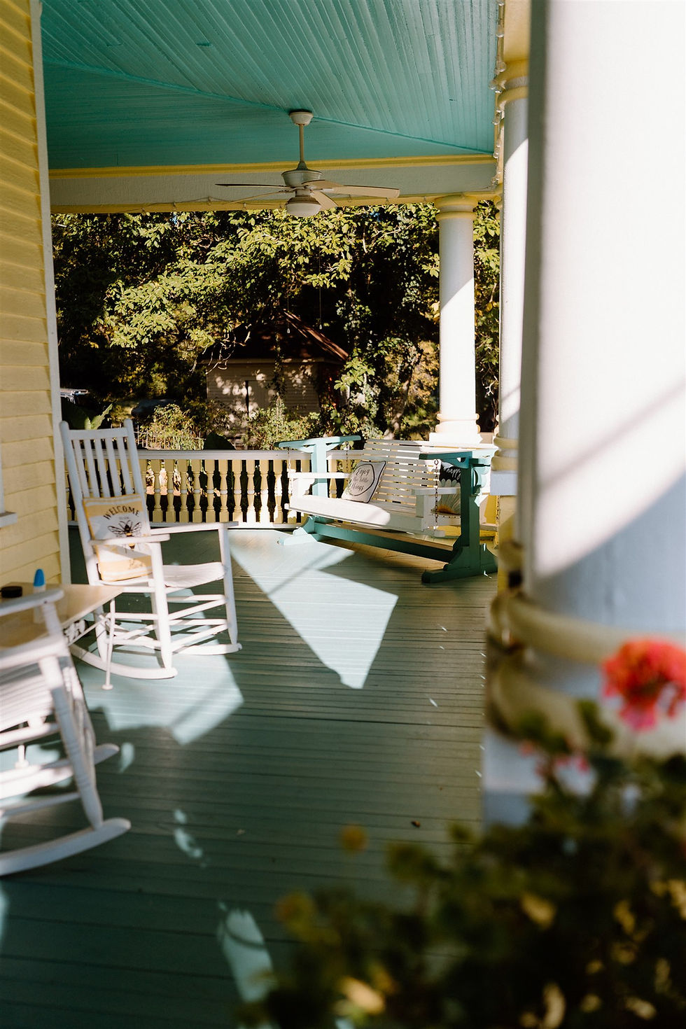 A sunlit porch with a white rocking chair and turquoise swing. A pillow reads "Welcome." Green trees are in the background.
