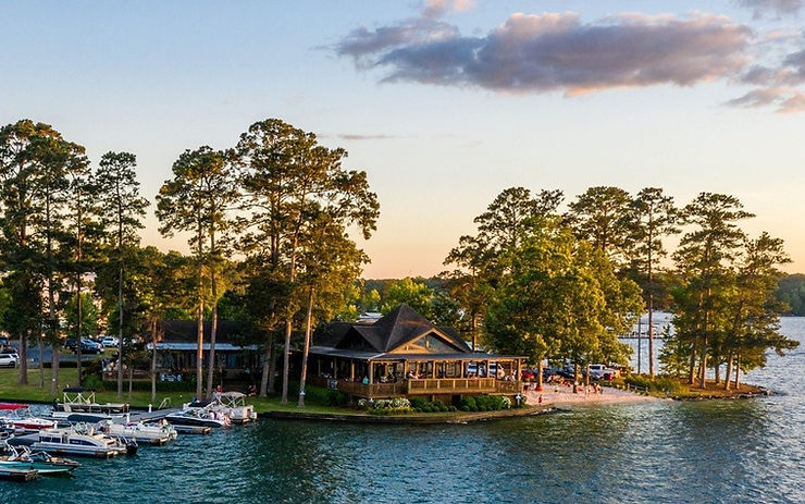 Lakeside restaurant surrounded by trees at sunset, with boats docked nearby. People gather on the patio, creating a relaxed, scenic ambiance.