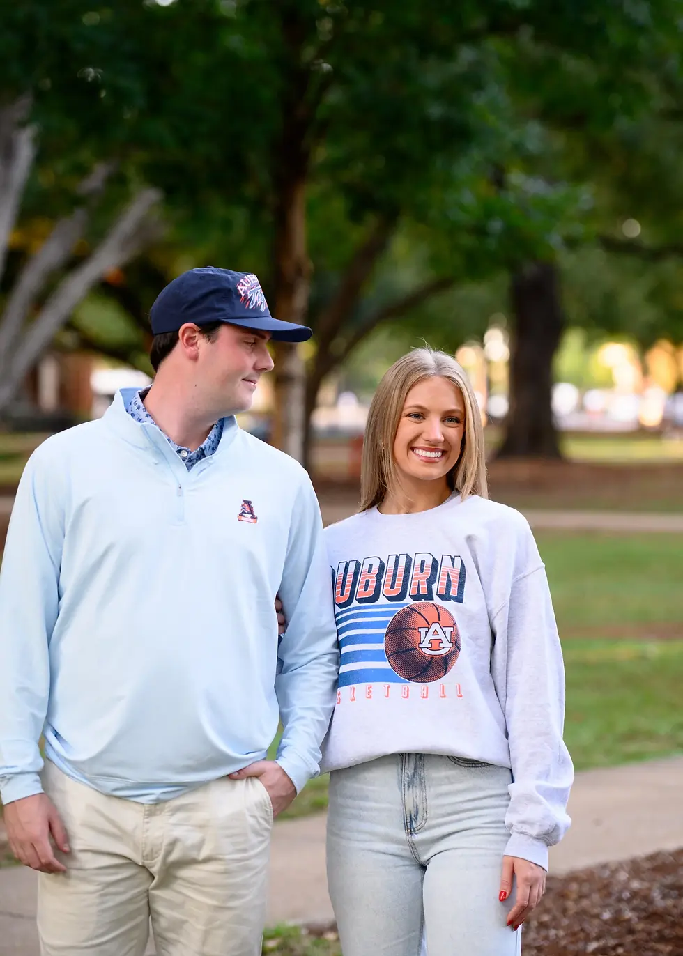 A smiling woman and a man stand arm-in-arm outdoors. She wears an Auburn sweatshirt; he wears a light blue pullover and cap. Lush trees behind.