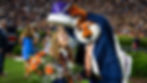 Mascot crowns a smiling girl holding flowers and a silver bowl, surrounded by people on a field. Crowd in the background, festive atmosphere.