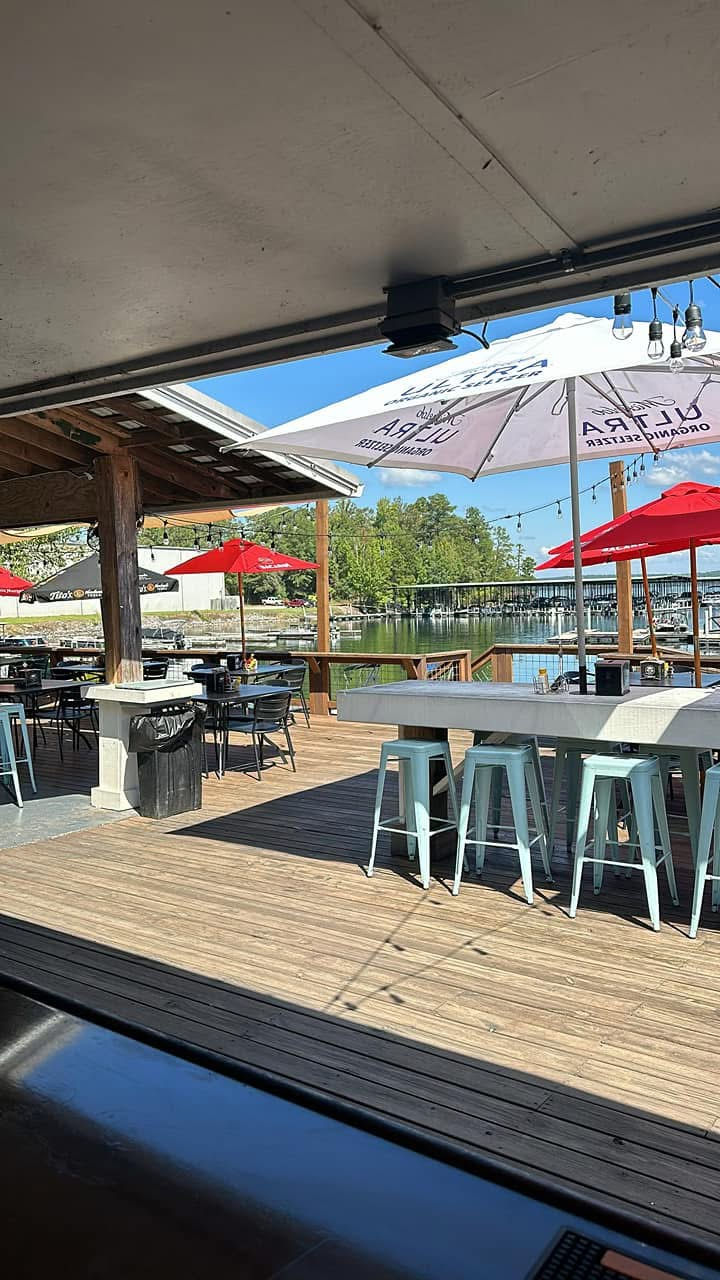 Outdoor lakeside patio with empty tables, stools, and red umbrellas. Midday sun casts clear shadows. Text on umbrella reads "Ultra."