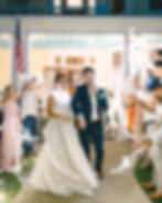 Bride and groom exit a house surrounded by guests with sparklers, smiling under string lights. American flag visible. Celebratory mood.