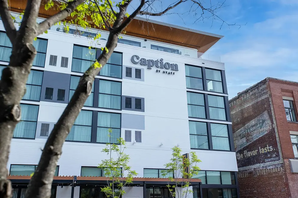 Exterior view of Caption by Hyatt Chattanooga Downtown at 105 W. Main St., a modern white multi-story hotel finished by Dream Team Finishes, a Chattanooga, Tennessee commercial painting company, photographed from street level through tree branches against a blue sky.