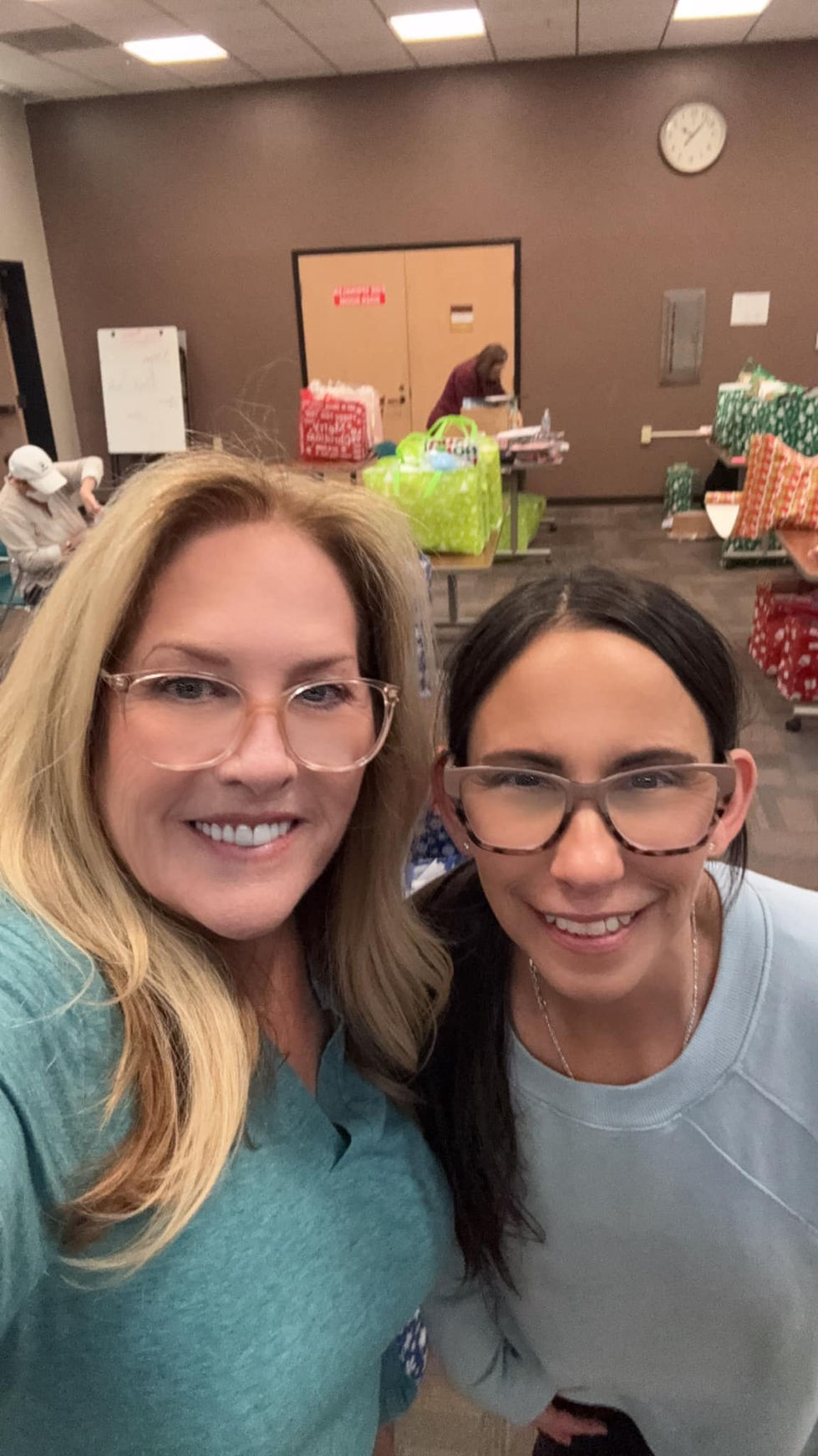 two woman with glasses pose smiling in front of tables full of wrapped gifts