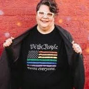 A white person with short peppery hair and white glasses stands in front of a brick wall with a black shirt on that says, "We the People means everyone".