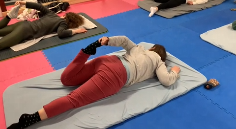 Katja auf dem Boden währen einer EML, sie erkundet ihren Fuß in Bauchlage hinter dem Rücken.