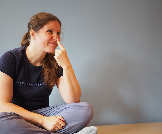 Katja Glock Neuro Horizons Practitioner sitzt im Schneidersitz auf ihrem Feldenkrais Tisch und tippt sich mit fragendem Gesichtsausdruck auf die Nase