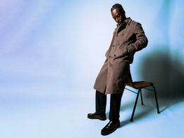 Kwabena Peprah seated in a studio wearing a long wool coat against a pale blue backdrop