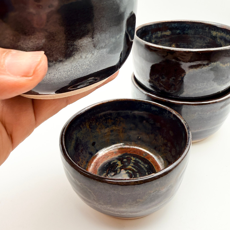 A hand holding one handmade ceramic bowl with a dark metallic tenmoku glaze with 3 bowls stacked on a white background.
