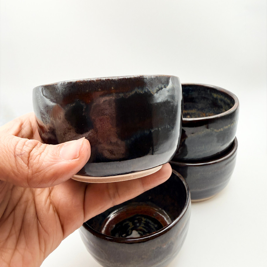 A hand holding one handmade ceramic bowl with a dark metallic tenmoku glaze with 3 bowls stacked on a white background.