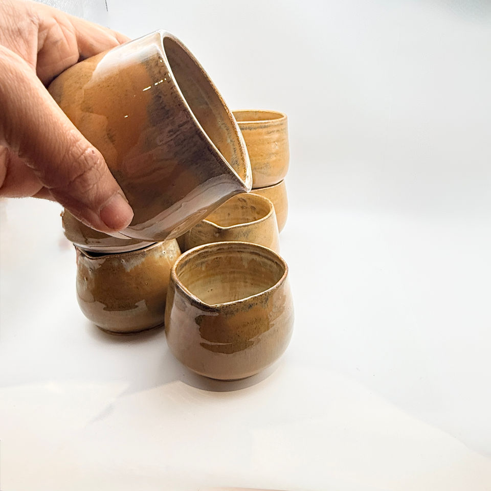 Hand holding a small handmade ceramic pitcher or small jugs with earthy ochre glaze, with more cups on a white background.