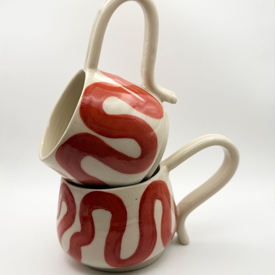Two hand-painted cream ceramic mugs with red wavy patterns stacked on a white background