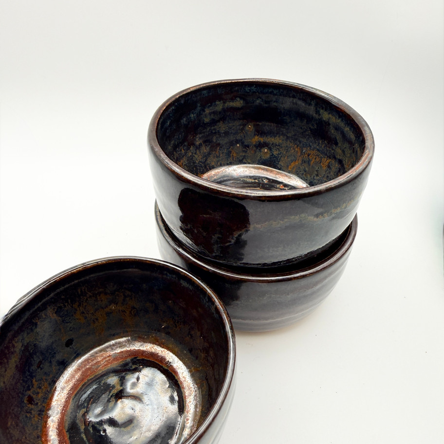 Three handmade ceramic bowls with a dark metallic tenmoku glaze stacked on a white background.
