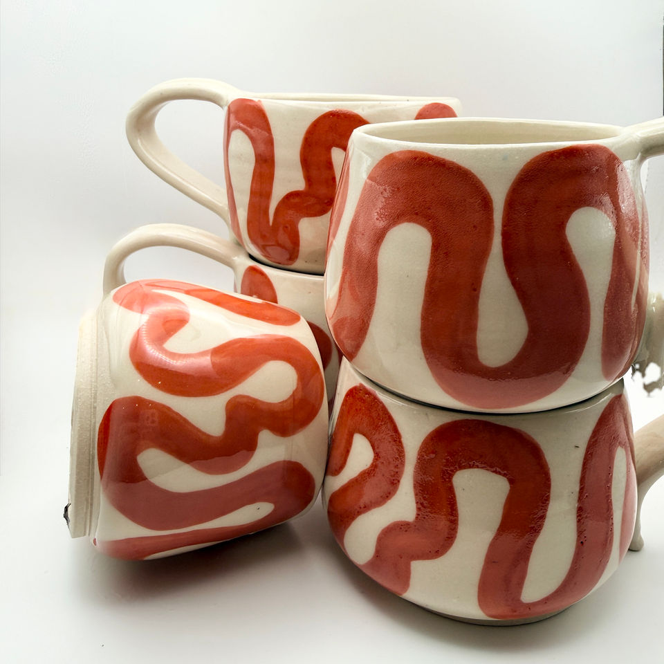 Four hand-painted cream ceramic mugs with red wavy patterns stacked on a white background