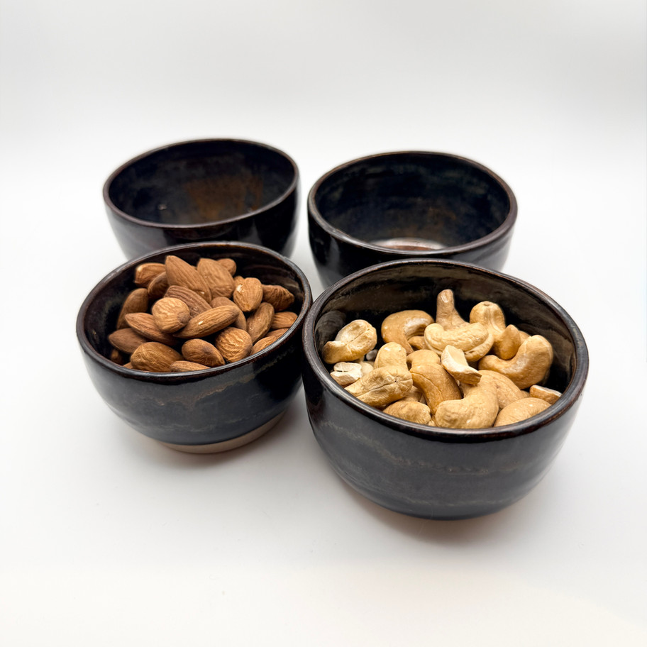 Dark ceramic bowls filled with raw almonds and cashews alongside two empty bowls on a white background.