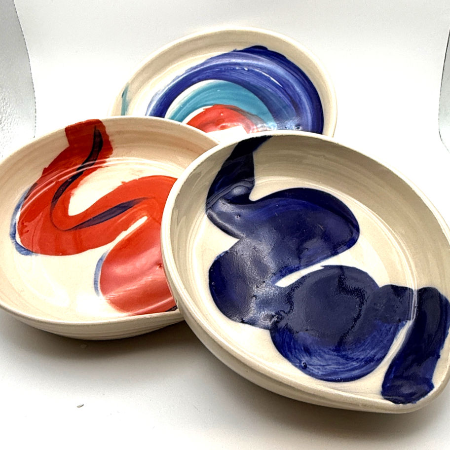 Three handmade cream ceramic spoon rests with blue and red abstract painted designs on a white background.