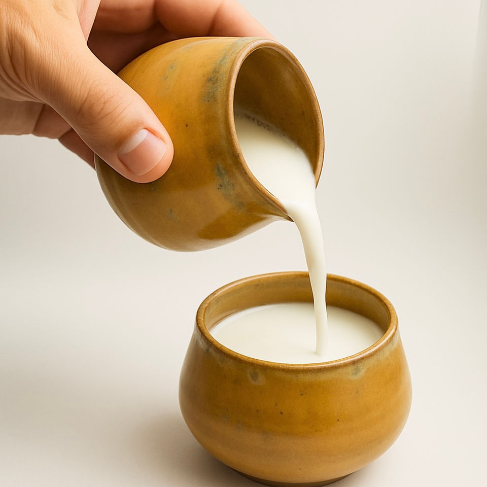 A hand pouring fresh white milk from an ocher ceramic pitcher into a matching cup against a plain background.
