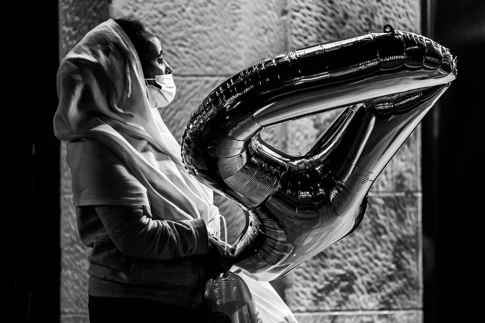 A woman stands against a stone wall at night, wearing a hooded scarf and a face mask, holding a large shiny balloon in the shape of the number four. Harsh side lighting reveals the texture of the balloon and the fabric around her, creating a dramatic black-and-white street scene that conveys waiting, hope, and quiet emotion in the city.