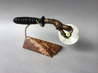 Pizza cutter, handmade: turned ebony handle with a chased copper replica of a lobster claw pinching a rotary cutting wheel; amboina burl stand. 1850