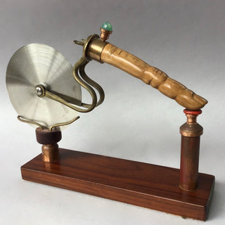 Handmade pizza cutter, carved olive wood handle shaped as finger with jewel ring; on mahogany stand with copper and glass fittings. 1925