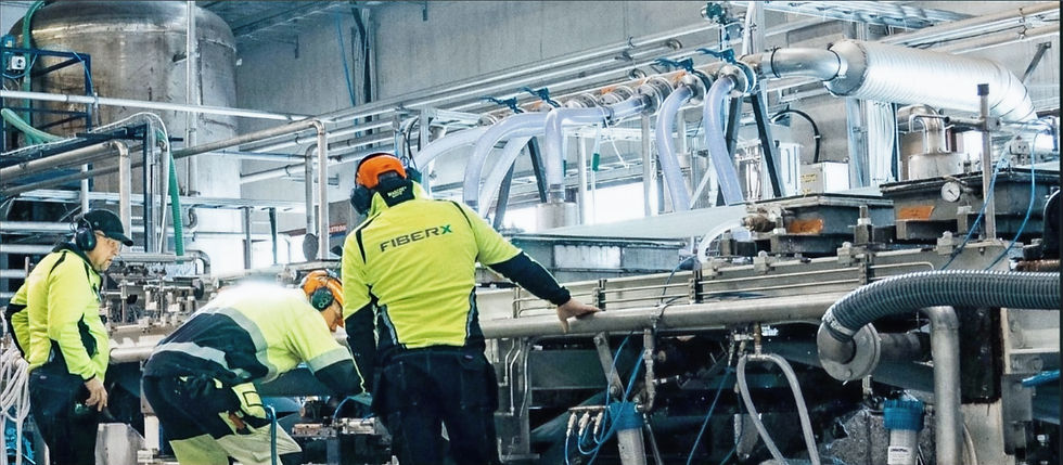 Three workers in bright safety gear inspect industrial machinery. Pipes and tubes fill the factory setting, with "FIBERX" on jackets.