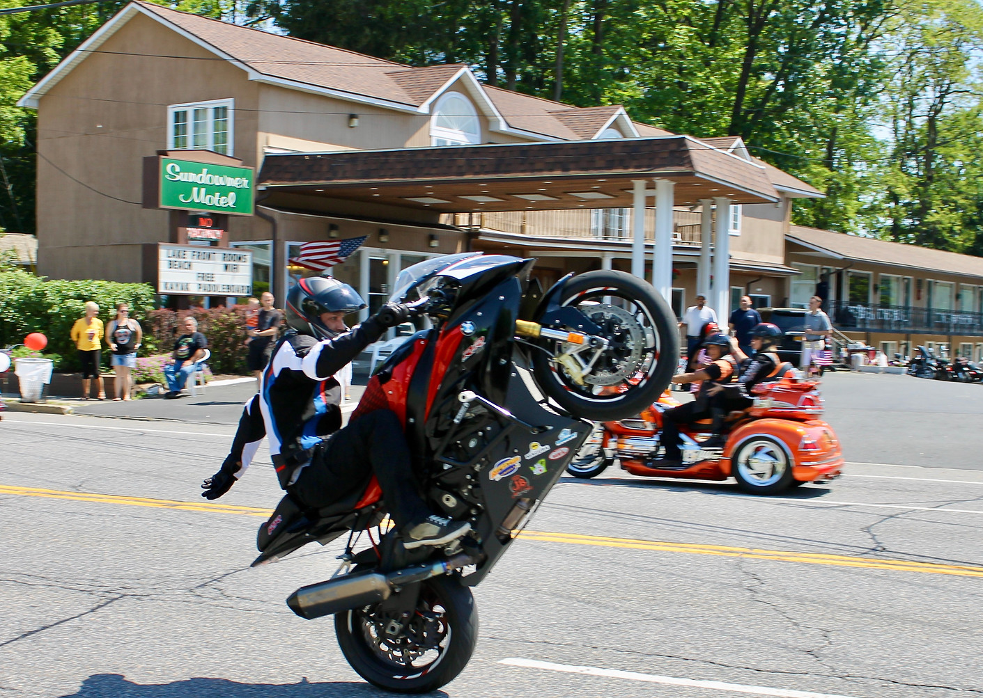 americade motorcyle rider popping a wheelie on canada street in lake george in front of the entrance to the sundowner on lake george with other americade motorcycles and spectators in the background