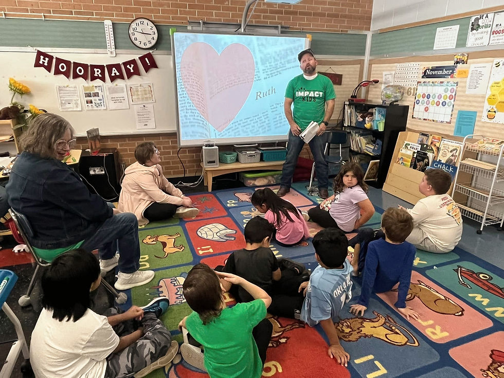 A teacher stands in a classroom with children seated on a colorful rug during an after school Bible club. A projection of a heart and "Ruth" is displayed.